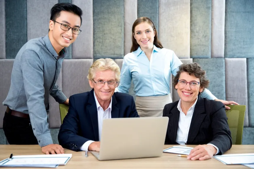 Smiling diverse team of four posing around a laptop in office environment as business directory lists your business